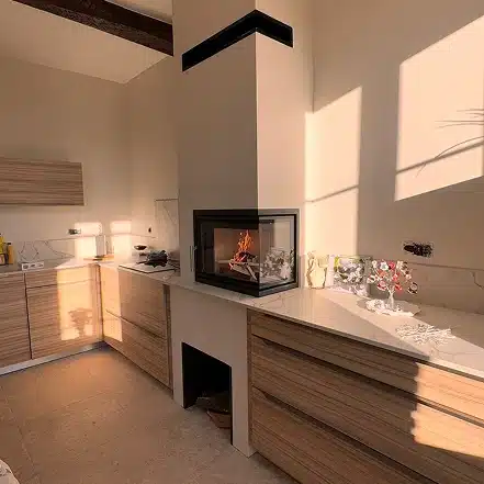 Cuisine moderne avec des armoires en bois clair, des comptoirs clairs et une cheminée encastrée au centre. La lumière du soleil projette des ombres chaudes sur les murs et le comptoir beige, avec des objets décoratifs exposés à côté de la cheminée.