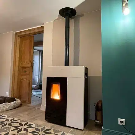 Un poêle à granulés moderne avec une flamme visible se trouve dans un coin d'une pièce au sol carrelé à motifs, près d'une porte en bois et d'un mur sarcelle avec un luminaire et une prise électrique.