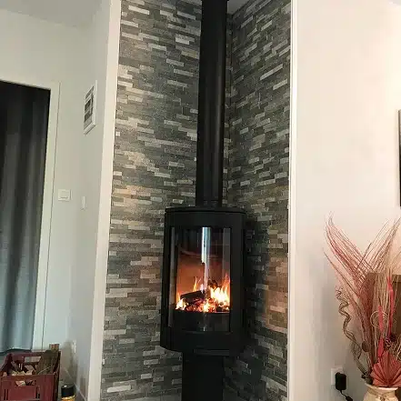 Un poêle à bois noir moderne avec une façade en verre est placé contre un mur d'angle avec des carreaux de pierre gris empilés. Les flammes sont visibles à l'intérieur et des plantes décoratives séchées se trouvent sur le côté droit.