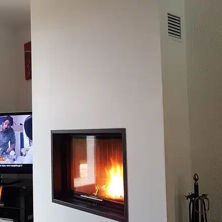 Une cheminée blanche moderne avec une façade en verre montre un feu vif à l'intérieur. À gauche, un téléviseur montre deux personnes discutant dans une cuisine. Un ensemble d'outils de cheminée en or se trouve à droite, contre un mur blanc.