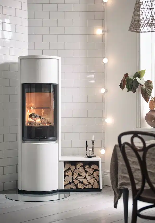 Un poêle à bois blanc moderne avec une porte vitrée montre un feu à l'intérieur. Le bois de chauffage est soigneusement empilé en dessous, et des guirlandes lumineuses sont suspendues aux murs carrelés blancs dans une pièce confortable et élégante avec une plante et une table de salle à manger à proximité.