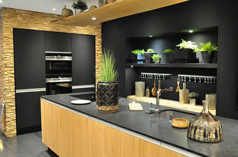 Cuisine moderne avec des murs noirs, des armoires en bois et des étagères ouvertes présentant des plantes en pot. Le comptoir accueille des vases décoratifs et un bol, et un four encastré est encastré dans un mur d'accent en pierre.