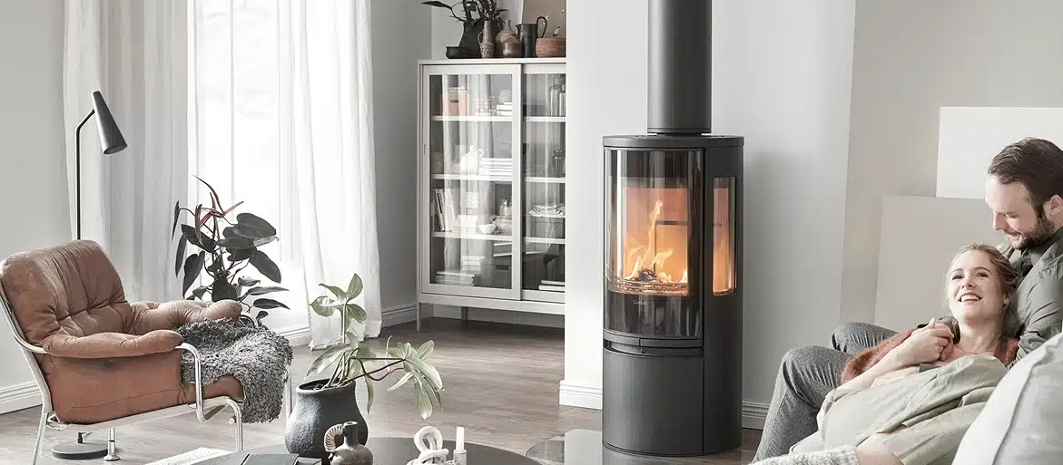 Un salon confortable avec un poêle moderne en bois noir, un fauteuil en cuir marron, des plantes d'intérieur et un couple qui se fait des câlins sur un canapé gris. La lumière chaude du poêle ajoute une ambiance réconfortante.