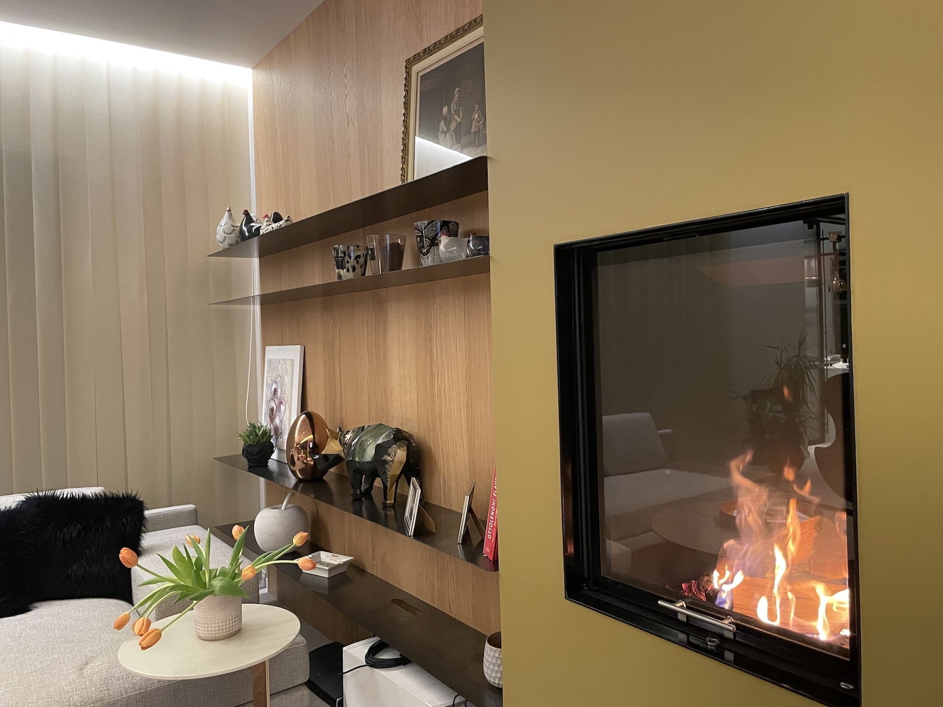 Un salon moderne avec des murs beiges, une cheminée sur la droite, des étagères flottantes avec du décor, et un vase de tulipes orange sur une petite table ronde à côté d'un canapé gris avec des coussins noirs et blancs.