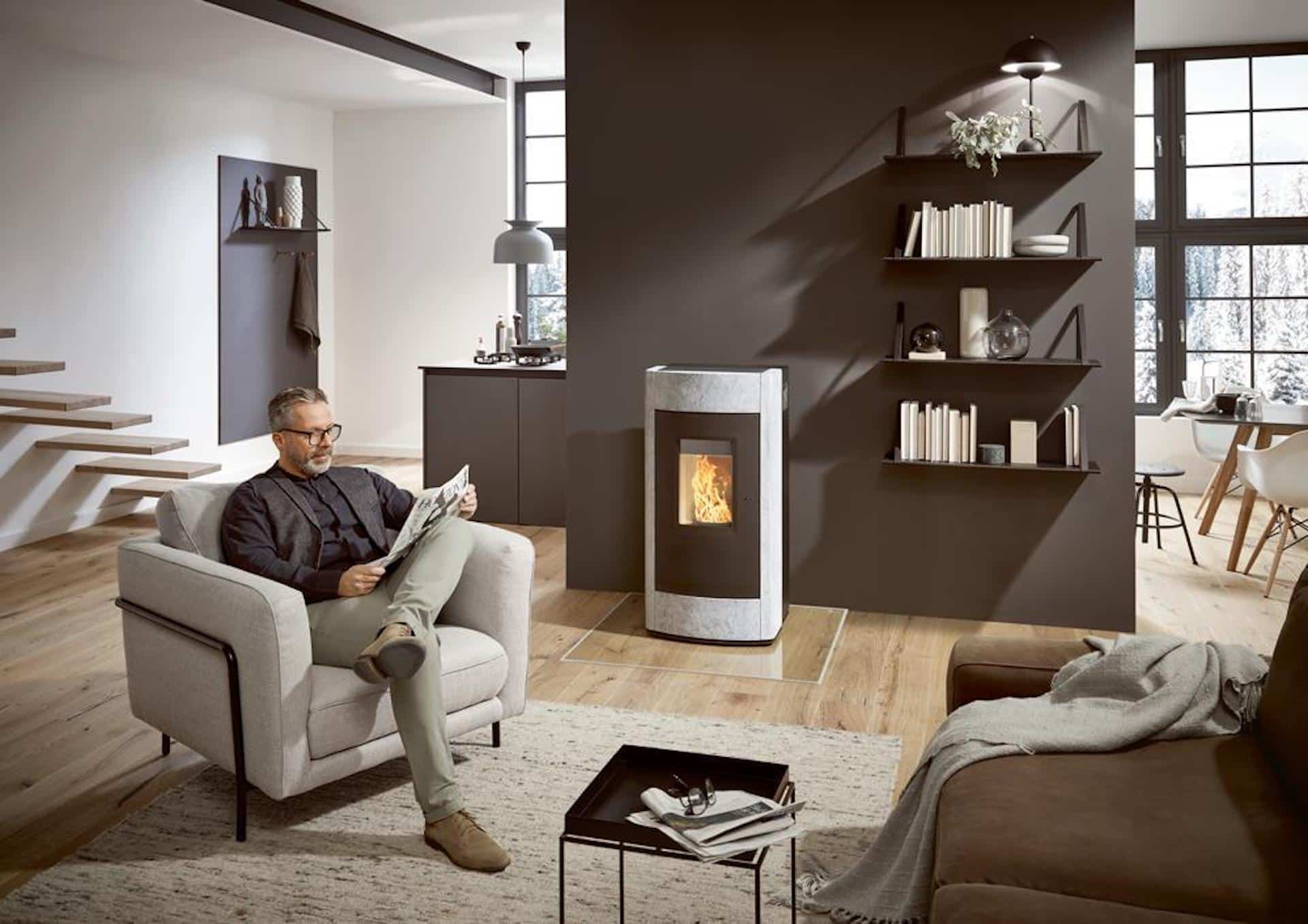 Un homme est assis dans un salon moderne et lit un journal. Il y a une cheminée allumée, des étagères murales avec des livres et de la décoration, et de grandes fenêtres qui laissent entrer la lumière naturelle. L'espace présente des tons neutres et un mobilier contemporain.
